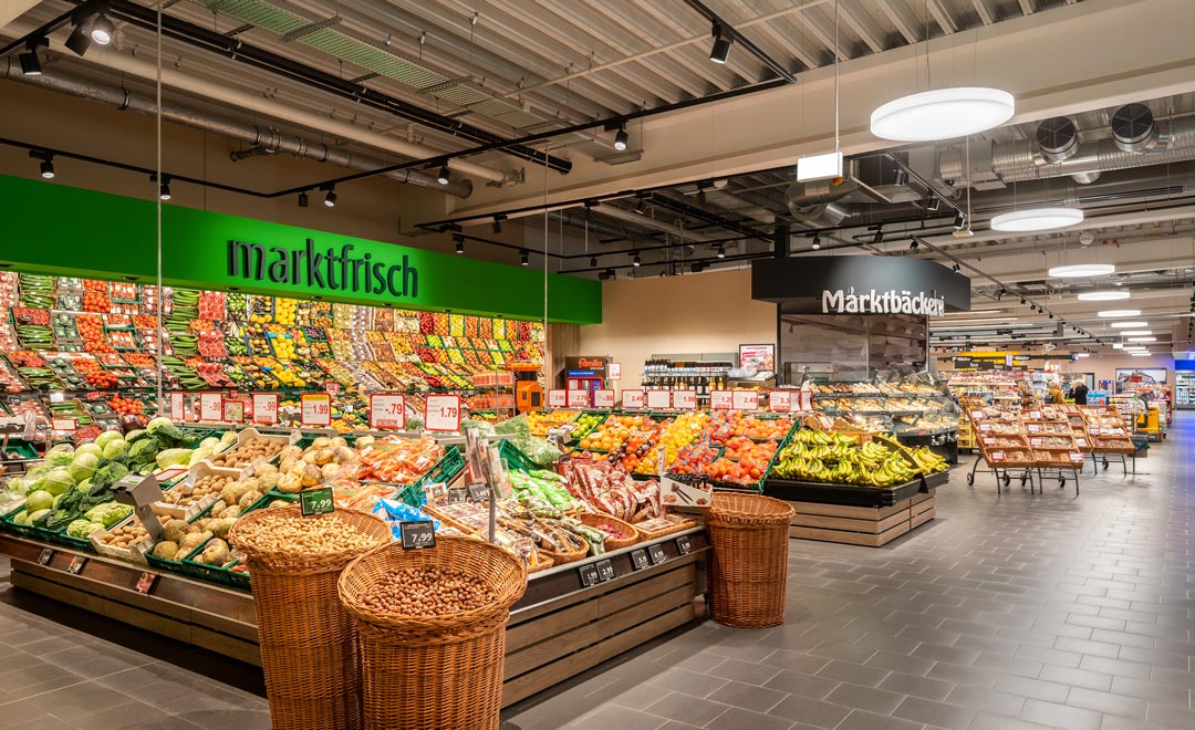 Sección de frutas y verduras con vistas hacia la entrada principal iluminada con el proyector B.Veo y la luminaria de suspensión Solegra. 